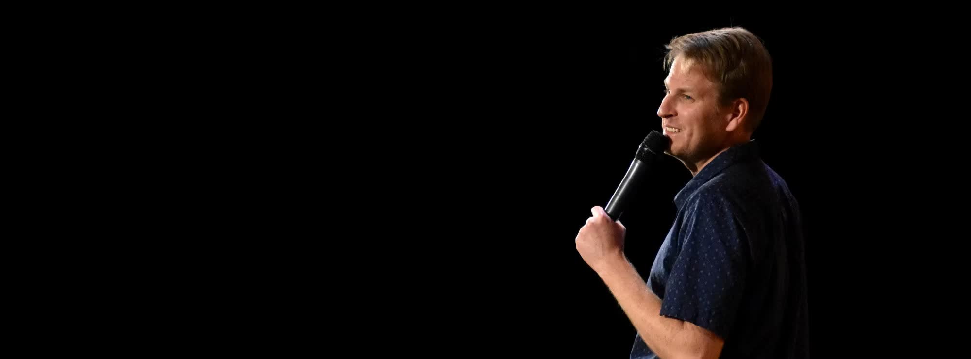 Mark Schumacher on a dark stage, smiling mid-joke with a microphone in hand