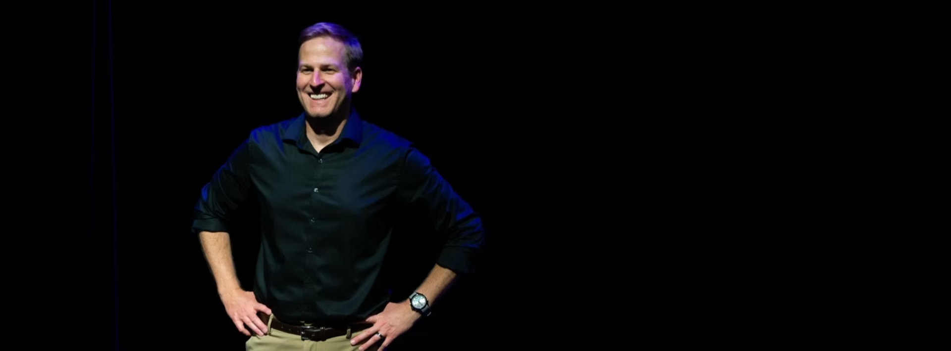 Mark Schumacher standing on stage under blue light, hands on hips, laughing with the audience